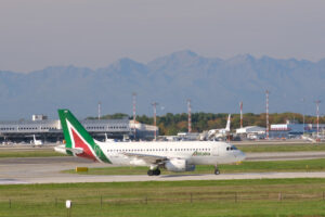 Alitalia Airbus A319-112  airplane taxiing on the Malpensa airport runway. In the background the buildings of  Cargo Terminal and parked airplanes. - MyVideoimage.com