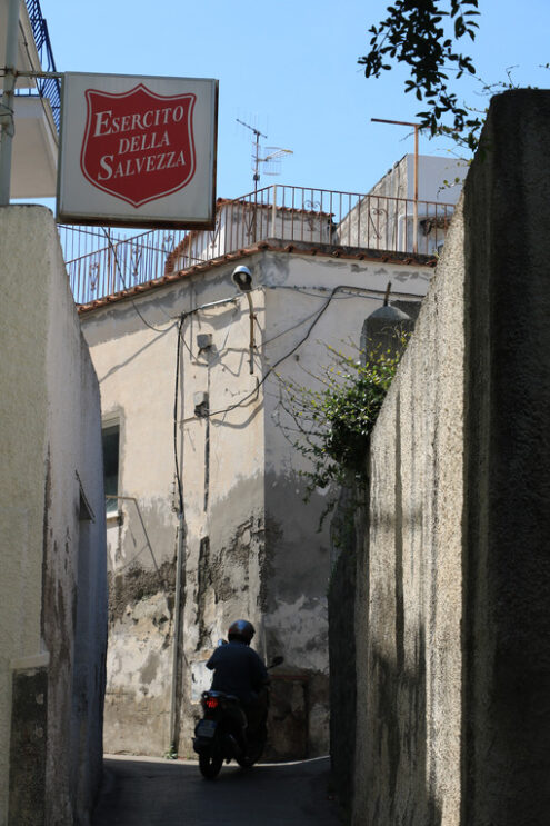 Alley with Mediterranean houses and high walls, sign of the Salvation Army. Person on a motorcycle. - MyVideoimage.com