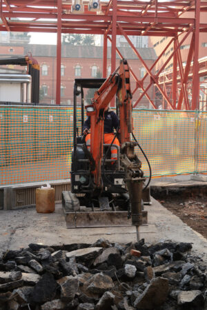 Asphalt demolition. Demolition of the asphalt of a road with a jackhammer. Small excavator with demolition hammer. In the background steel trellis with plant pipes. - MyVideoimage.com | Foto stock & Video footage