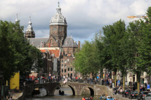 Bridge on a city canal with people walking. In the background an ancient church with a dome and bell tower. - MyVideoimage.com