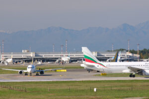 Bulgaria Air Embraer 190/195 airplane on the Malpensa airport runway. In the background the buildings of Terminal 1 and parked airplanes. - MyVideoimage.com