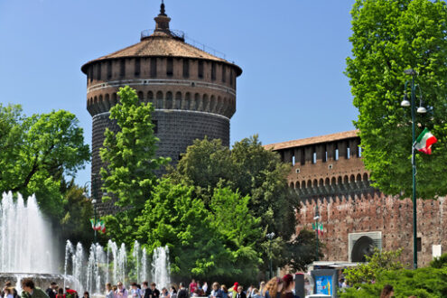 Castello Milano. Sforza Castle in Milan. The red brick walls and the tower. Many people walk near the Castle. In the foreground the fountain and the trees. - MyVideoimage.com | Foto stock & Video footage