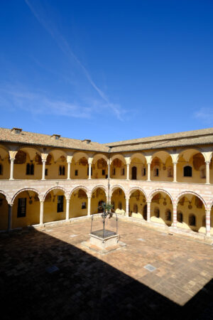 Cloister whit columns in Assisi. Cloister with columns and arches at the Basilica of San Francesco in Assisi. In the center of the courtyard there is a well in beige stone. - MyVideoimage.com | Foto stock & Video footage