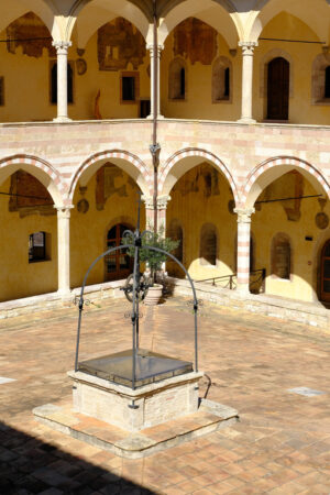 Cloister with columns and arches at the Basilica of San Francesco in Assisi. In the center of the courtyard there is a well in beige stone. - MyVideoimage.com