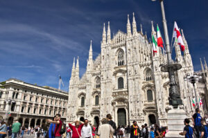 Duomo Milano. Milan Cathedral facade with flags on blue sky. The facade of the cathedral with many people walking. Flags waving on the blue sky. - MyVideoimage.com | Foto stock & Video footage