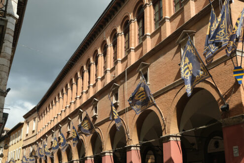 Foligno Umbria. Strada del paese. Main street of Foligno with waving flags on the facades of the houses. The ancient palaces lit by the sun with cloudy sky. - MyVideoimage.com | Foto stock & Video footage