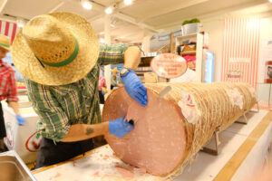 Large mortadella salami from Bologna. A pork butcher with a cowboy hat cuts a big salami. - MyVideoimage.com