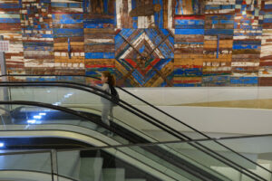 Little girl on an escalator at Bari airport. Decorative panel with wooden slats made from old boats. - MyVideoimage.com