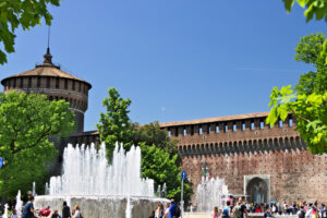 Sforza Castle in Milan. The red brick walls and the tower. Many people walk near the Castle. In the foreground the fountain and the trees. - MyVideoimage.com | Foto stock & Video footage