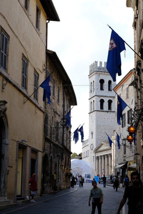 Street in the city of Assisi with the background of the civic tower and the temple of Minerva. Flags and people in the foreground. - MyVideoimage.com | Foto stock & Video footage