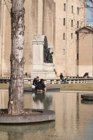 Two people sitting on the edge of a pond. Poplar trees in a tub of water. In the background the Palazzo della Pilotta in Parma. - MyVideoimage.com