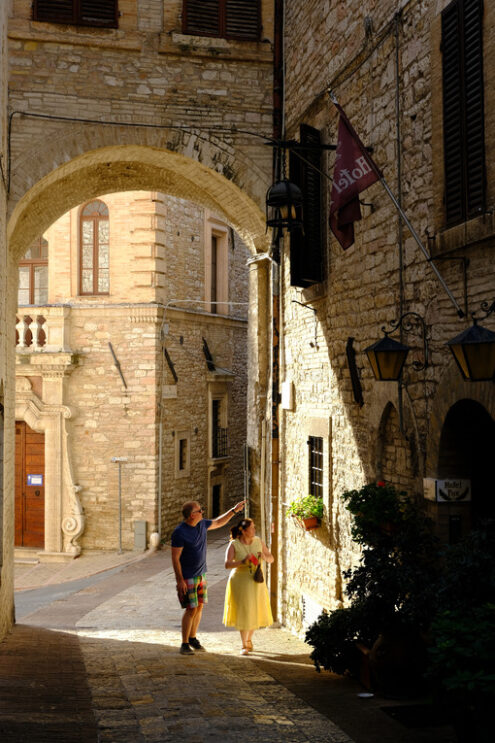 Two tourists walk in an ancient street under an arch in the city of Assisi. The sun illuminates the houses. - MyVideoimage.com | Foto stock & Video footage