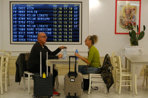 Two travelers sitting at a bar table in Bari airport. On the bottom, the board with the timetable for departing flights. - MyVideoimage.com