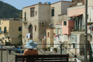 Woman with hat sitting on a bench. In the background old houses - MyVideoimage.com